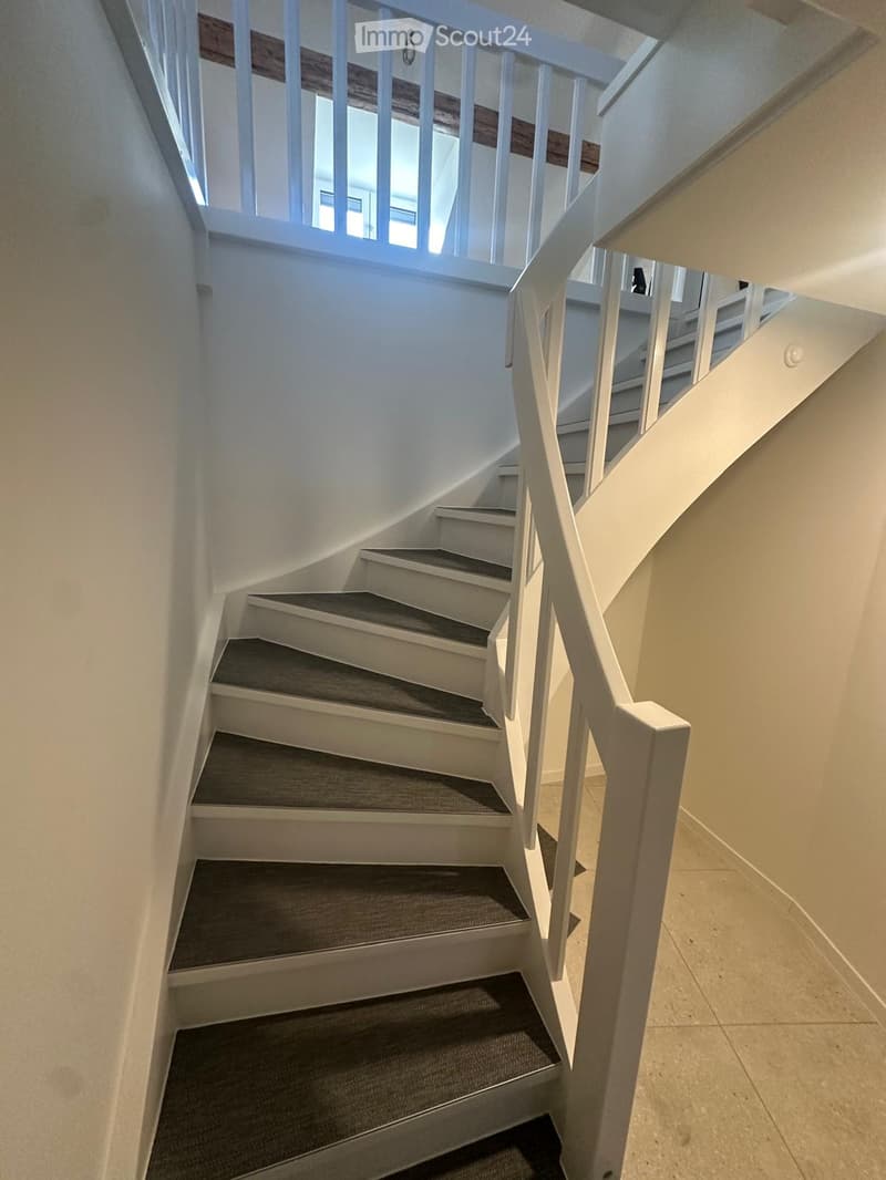 Spiral staircase with white wooden railings and gray carpeted steps, leading to an upper level with visible windows and light-colored walls.