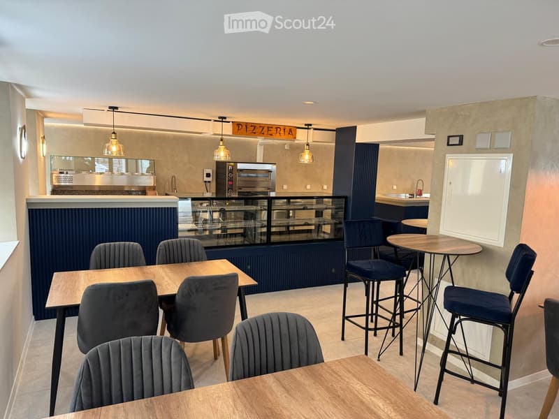Modern pizzeria interior with wooden tables, upholstered chairs, bar stools, and a visible kitchen area with an oven and a sink.
