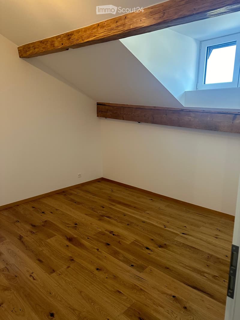 Empty room with wooden flooring, white walls, and wooden beams on the ceiling. A small window is visible on the right side.