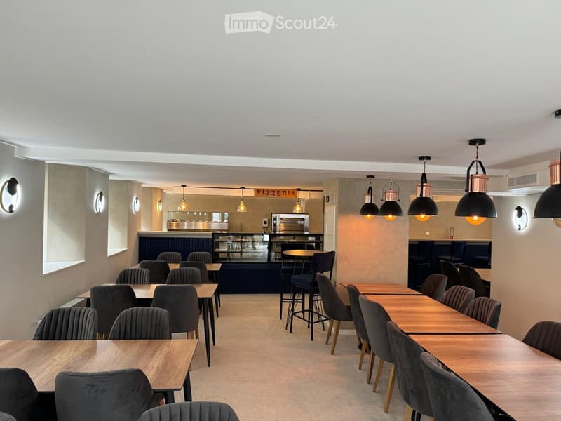Modern restaurant interior with wooden tables, grey upholstered chairs, pendant lights, and a visible kitchen area with an oven.
