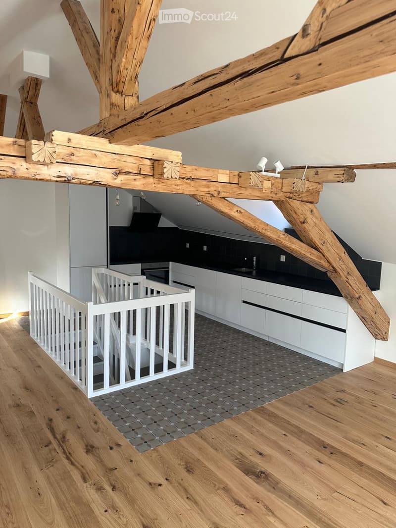 Open-plan living and kitchen area with exposed wooden beams, modern white kitchen cabinets, and a grey tiled floor.