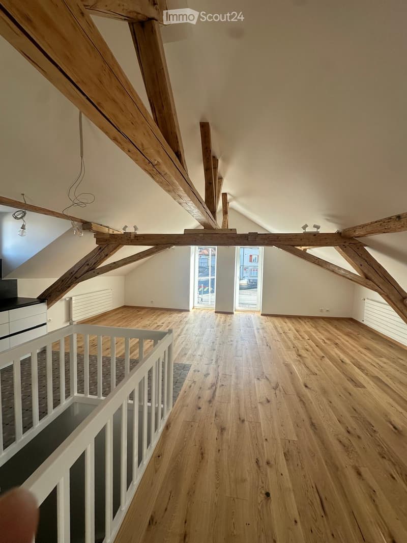 Empty attic space with wooden beams, wooden flooring, and large windows offering a view of the street.