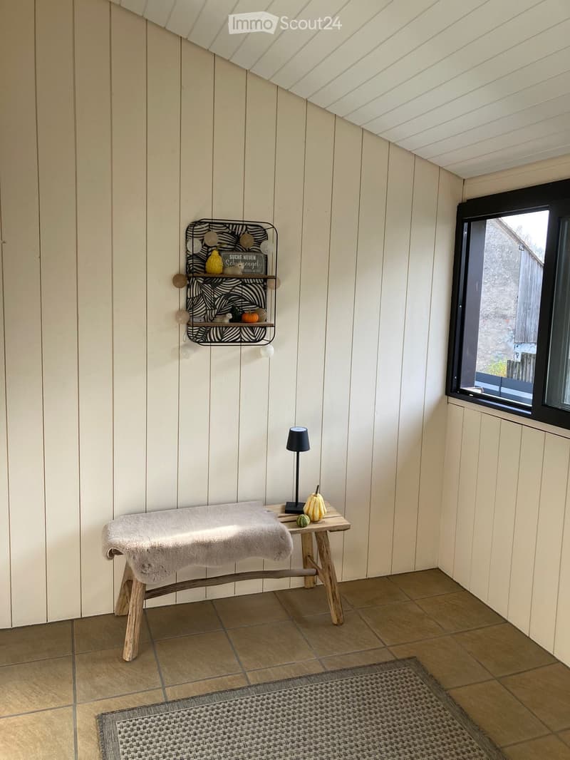 Cozy, slanted-ceiling room with wooden walls, tiled floor, small wooden table with lamp, stool with towel, shelf with decorative items, and woven rug.