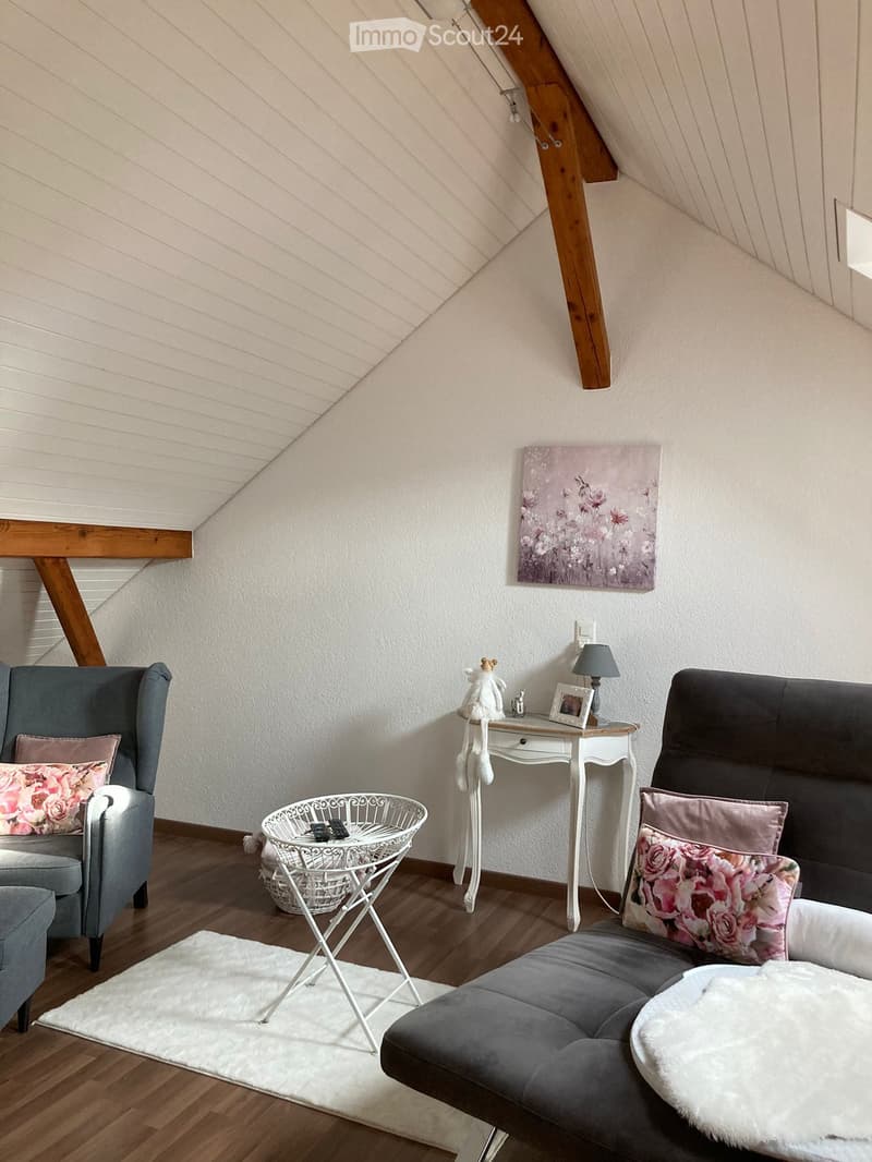Cozy attic living room with slanted wooden ceiling, grey sofa, armchair, small white table, lamp, and decorative items.