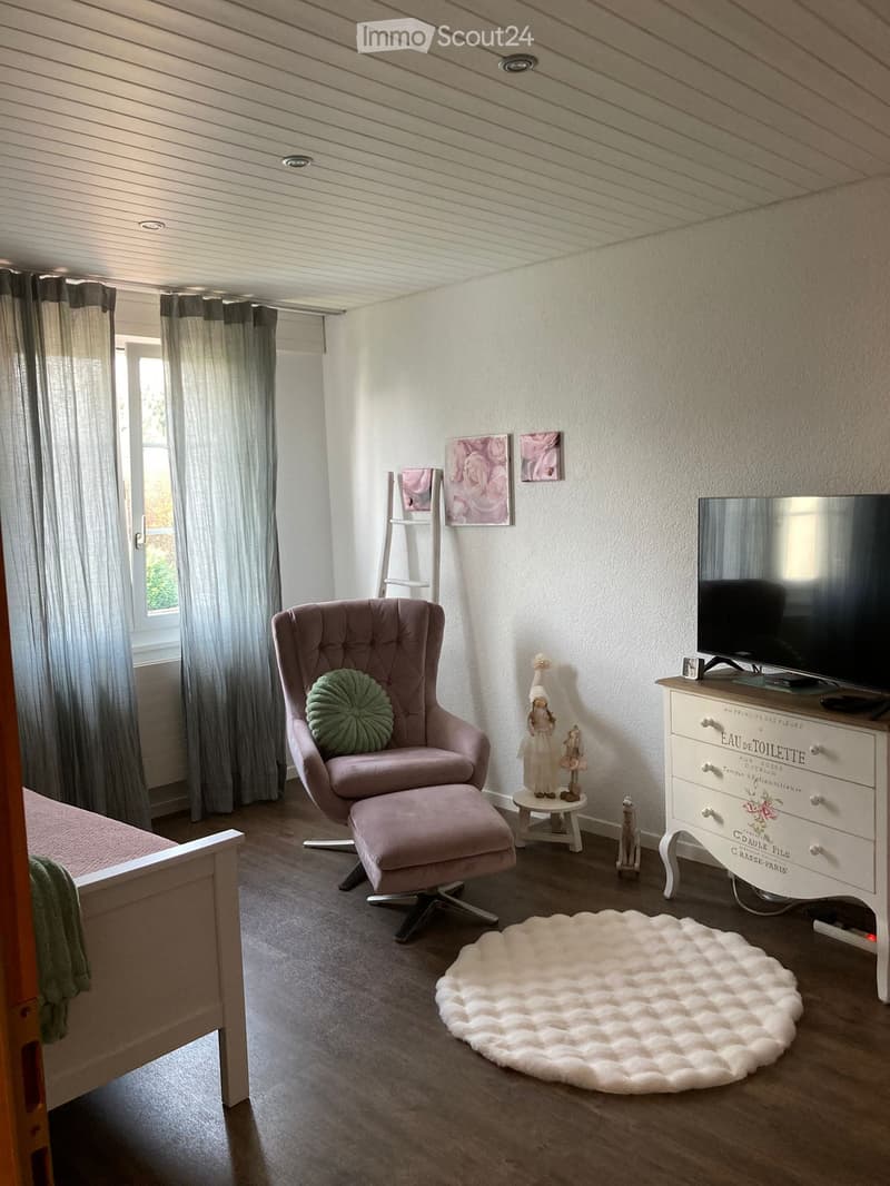 Cozy room with a pink armchair, matching ottoman, white shaggy rug, TV on a white cabinet, small table with decorative items, and a bed visible on the left side.