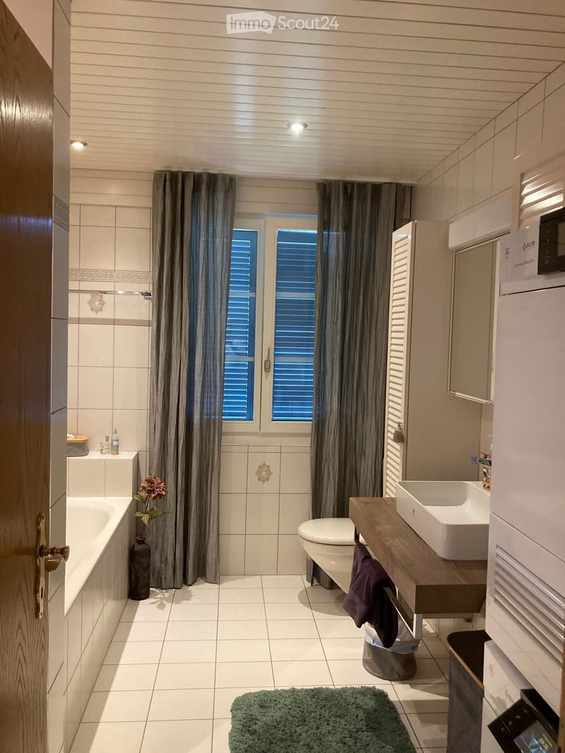 Modern bathroom with white tiles, a bathtub, toilet, sink, and a wooden vanity. Features include a large window with blinds, a decorative vase with flowers, and a green bath mat.