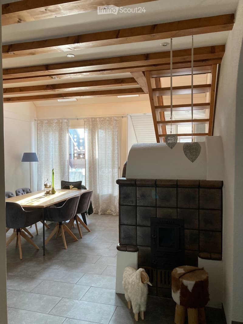 Dining area with a wooden table, chairs, and a fireplace with a decorative stool in front. There is a lamp, a vase on the table, and curtains on the windows.