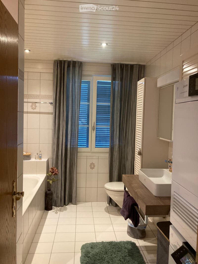 Modern bathroom with white tiles, a bathtub, toilet, sink, and a large window with curtains. Includes a wooden vanity, a mirror, a decorative vase, and a green rug.
