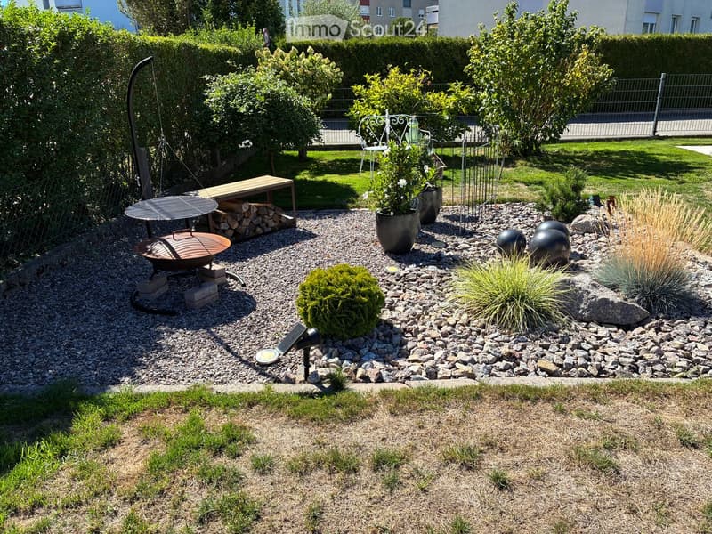 Well-maintained garden with gravel pathways, decorative plants, a fire pit, a swing, and a bench. Features include spherical sculptures, potted plants, and a stack of firewood.
