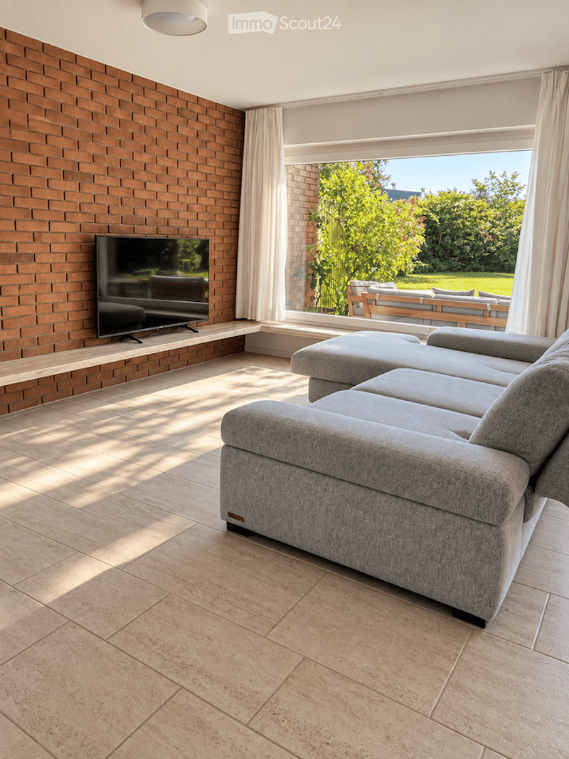 Modern living room with a grey sectional sofa, ottoman, flat-screen TV on a brick wall, large windows with white curtains, and a view of a garden.