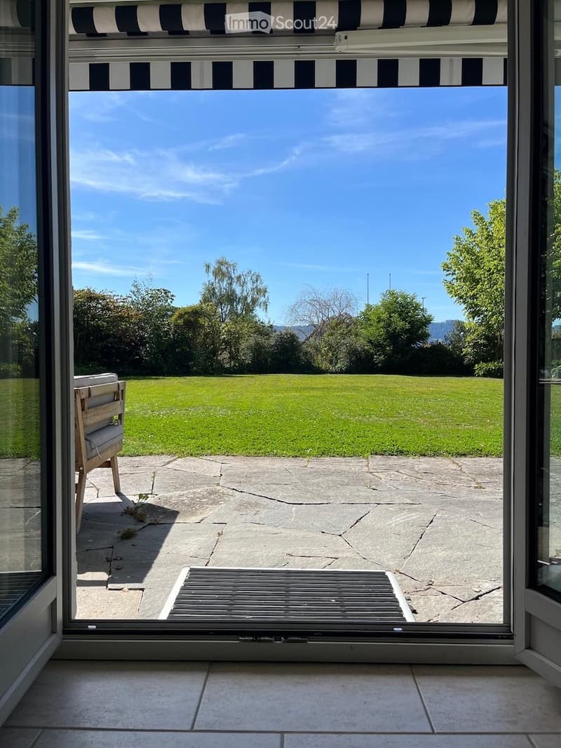 View from inside a house onto a well-maintained garden with a lawn, trees, and a clear blue sky. A wooden chair is visible on the patio.