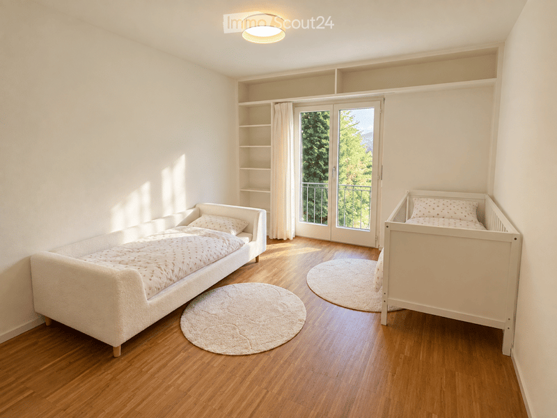 Bright bedroom with a sofa bed, a crib, round rugs, built-in shelves, and a large window with a view of greenery leading to a balcony.