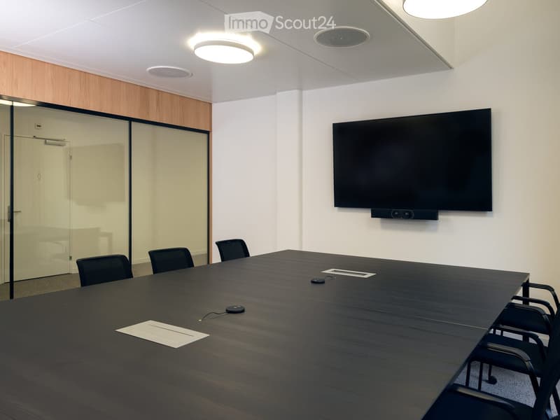 Modern conference room with a long dark wooden table, several black chairs, a large flat-screen TV mounted on the wall, and a glass partition with a door.