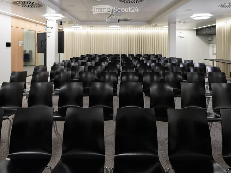 Modern conference room with rows of black chairs, white walls, and a projector screen at the front.