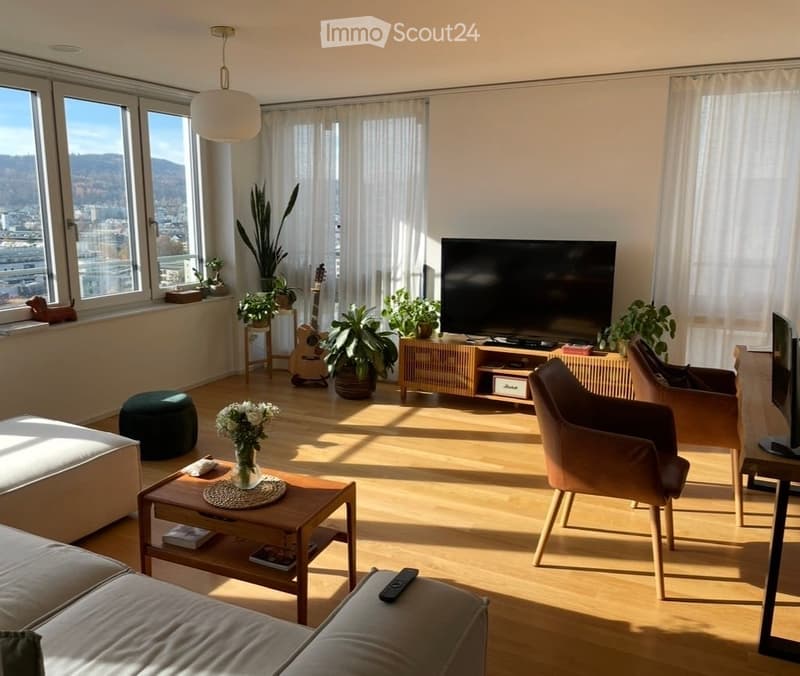 Bright living room with large windows offering a city view, featuring a grey sectional sofa, wooden coffee table, armchairs, a guitar, and several potted plants.