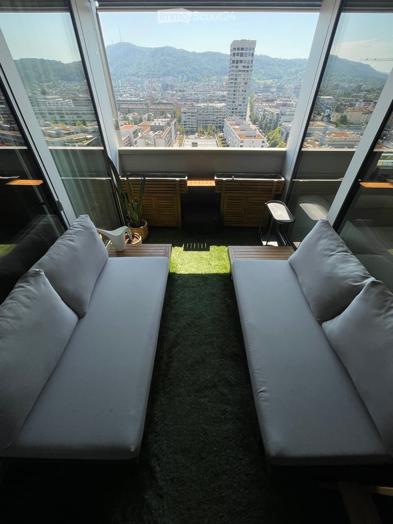 Modern balcony with two sofas, small tables, and a potted plant, offering a panoramic city view.