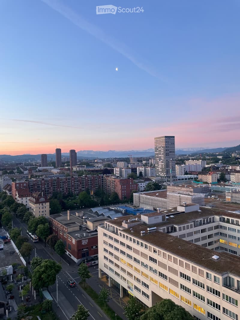 Aerial view of a cityscape at sunset, featuring a mix of residential and commercial buildings, with visible streets, trees, and distant mountains.