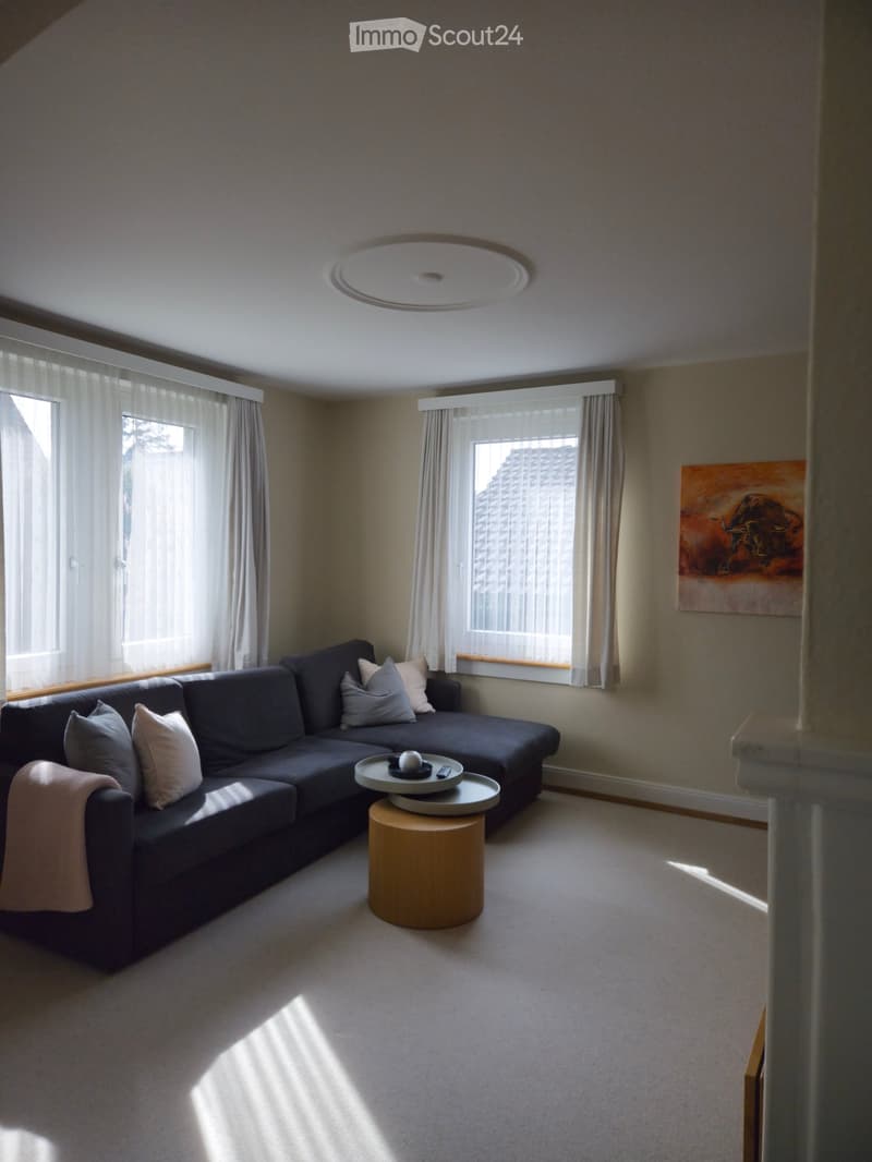 Modern living room with a grey sectional sofa, wooden coffee table, decorative pillows, and a painting on the wall. Large windows with white curtains allow natural light to fill the space.