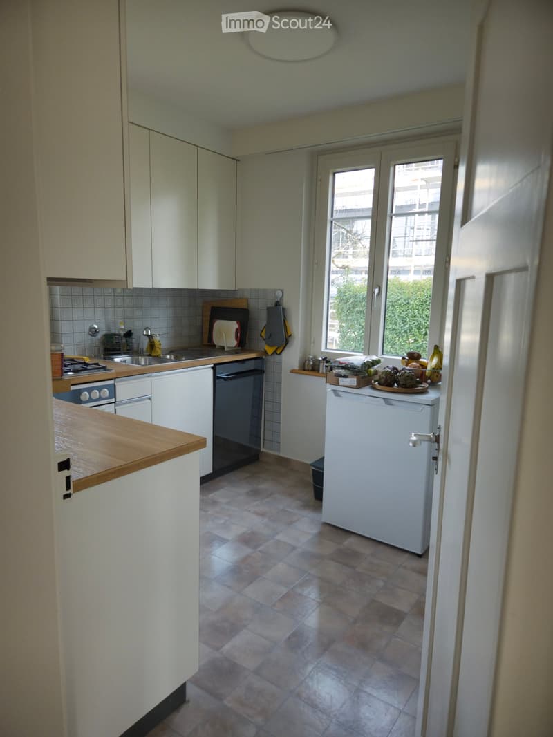 The kitchen features white cabinets, a wooden countertop, a tiled backsplash, and a tiled floor. Visible appliances include a refrigerator, oven, and sink. A cutting board and oven mitt are placed on the counter, along with a dish rack and a few kitchen i