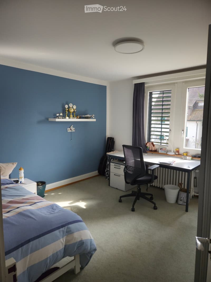 Bedroom with a bed, desk, chair, shelf with trophies, guitar, and window with curtains.