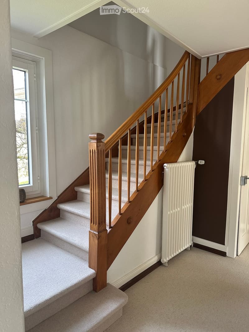 Spacious hallway with a wooden staircase featuring a polished handrail and balusters, carpeted stairs, a white radiator, and a window letting in natural light.