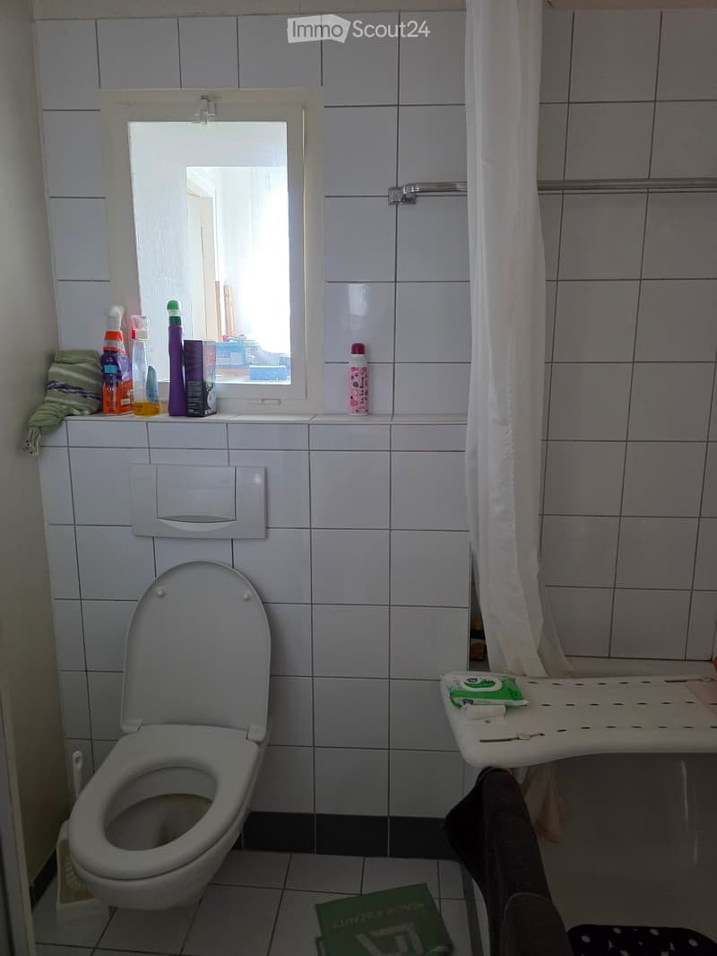 A small bathroom with white tiled walls and floor, featuring a toilet, a mirror above a sink, a shower curtain, and various cleaning supplies on the windowsill.