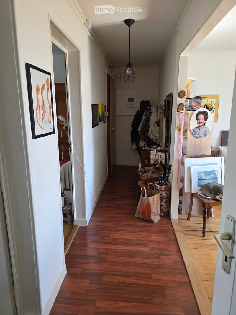 A narrow hallway with wooden flooring, white walls, and several doors. There are various items including artwork, a coat rack, a small table, and a hanging light fixture.