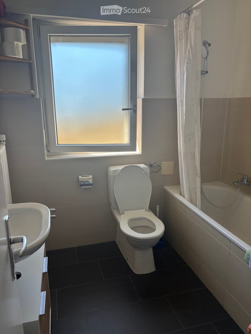 Modern bathroom with a toilet, bathtub, sink, and shower curtain. Features a window with frosted glass and a radiator below it.