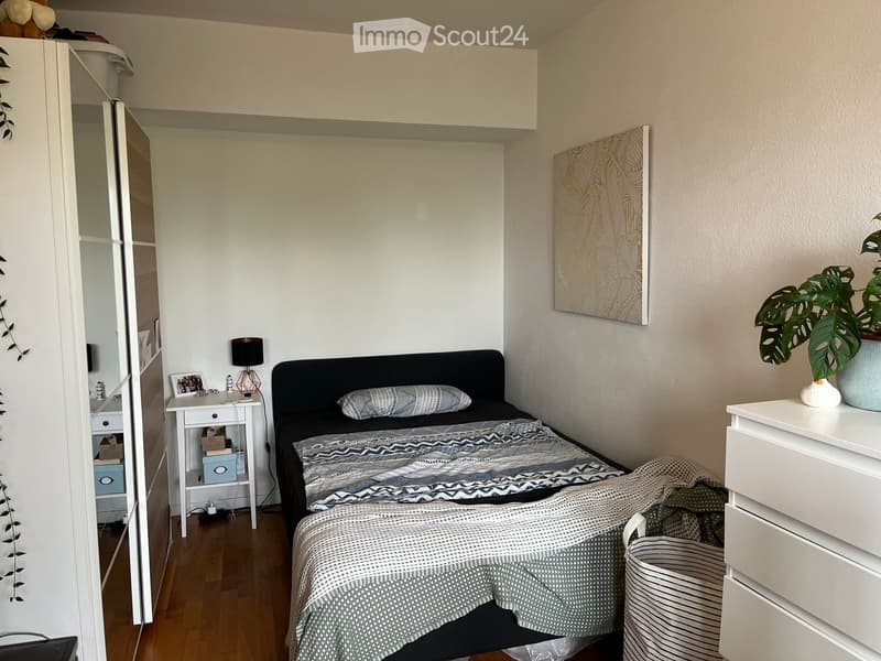 A modern, well-lit bedroom with a double bed, nightstand, lamp, and decorative plant. The room features a sleek design with a mix of white and dark tones.