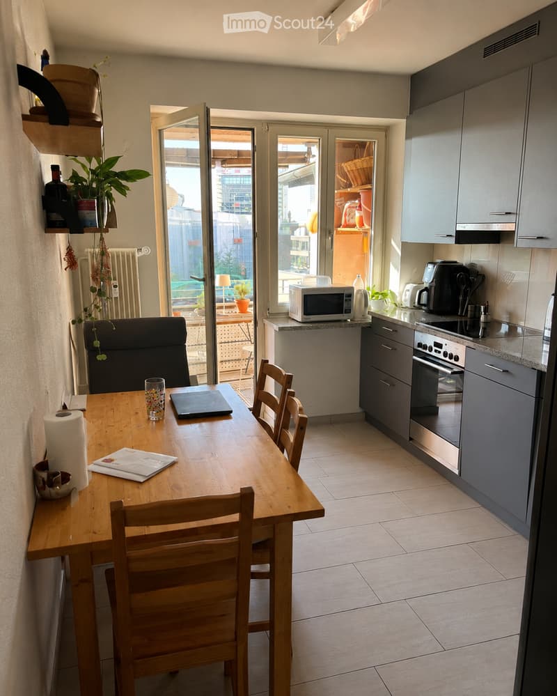 A modern kitchen and dining area with a wooden dining table, four chairs, a laptop, a microwave, an oven, and a coffee maker. The space features grey cabinets, a tiled floor, and a large window with a view of the city.