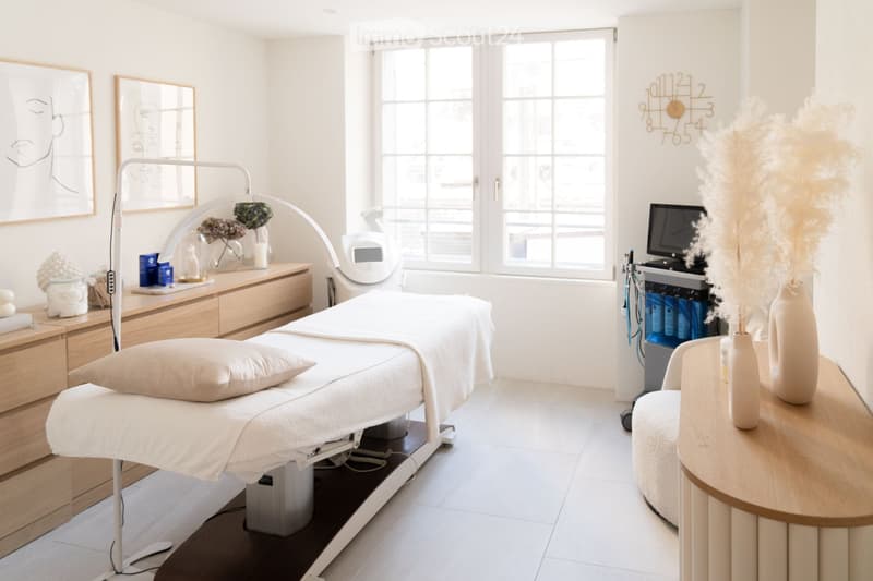 A modern, well-lit treatment room with a treatment table, a chair, a cabinet, a vase with dried flowers, a screen, and a clock on the wall.