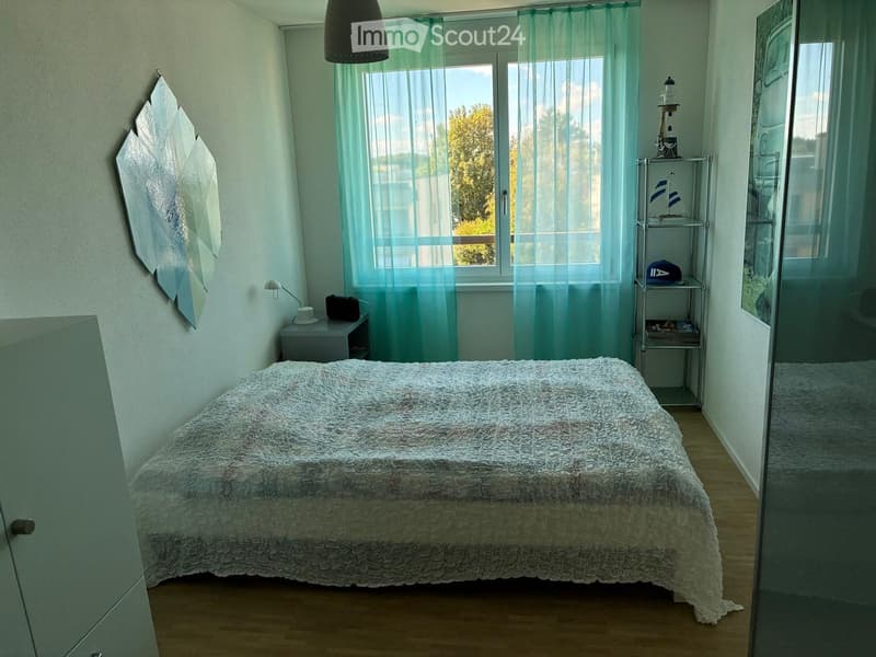 Cozy bedroom with a large bed, nightstand, wall-mounted lamp, decorative mirror, and a small shelf with various items.