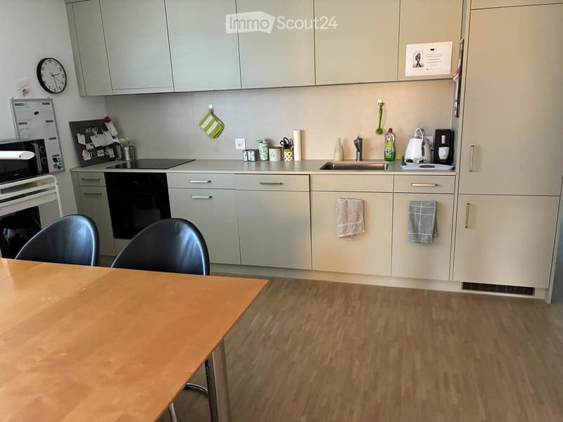 Modern kitchen with light grey cabinets, wooden floor, and stainless steel appliances. Includes a table with two chairs, a sink, coffee maker, and various kitchen items on the counter.