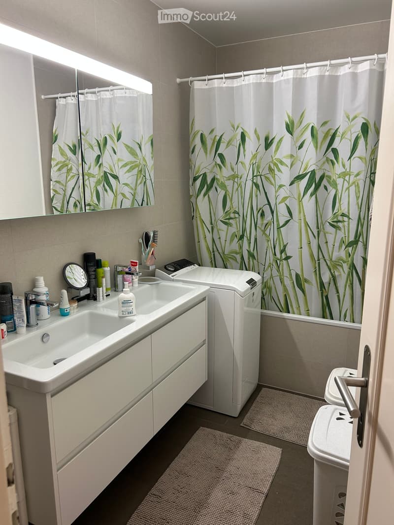 Modern bathroom with a double sink, mirror, toilet, bathtub with bamboo-patterned shower curtain, and various toiletries on the sink counter.