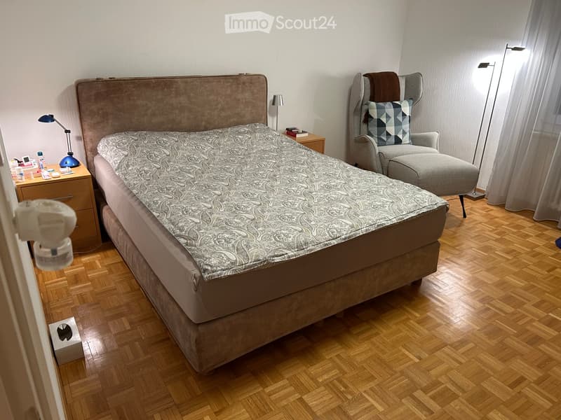 A well-furnished bedroom with a large bed, nightstand, armchair, and a lamp. The room features wooden flooring and large windows with curtains.