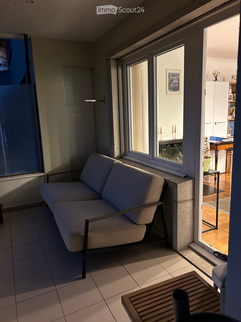Modern living room with a grey sofa, wooden coffee table, large windows, and a view into an adjacent room with a desk and shelves.
