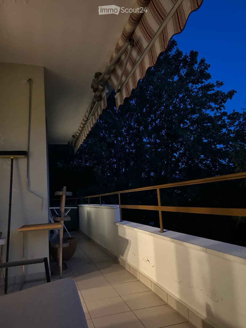Balcony with tiled floor, white walls, and a striped awning, featuring a railing and a view of trees.