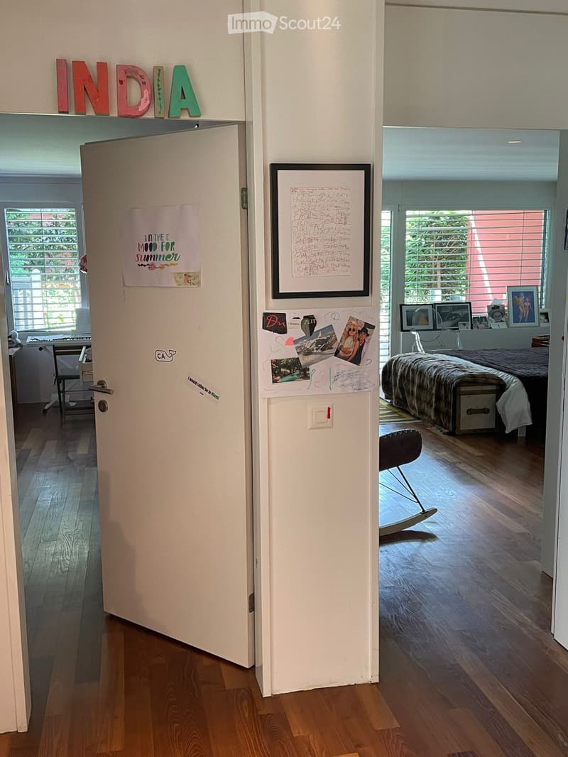 The door is open, wooden floor, white walls, bed in the room, pillows on the bed, some pictures on the wall, and a rocking chair.