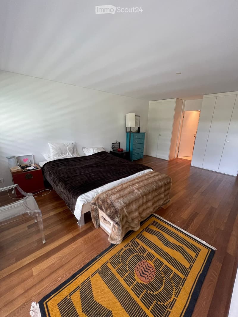 Wooden floor, bed with fur blanket and pillows, nightstand with items, glass desk, blue cabinet, closets