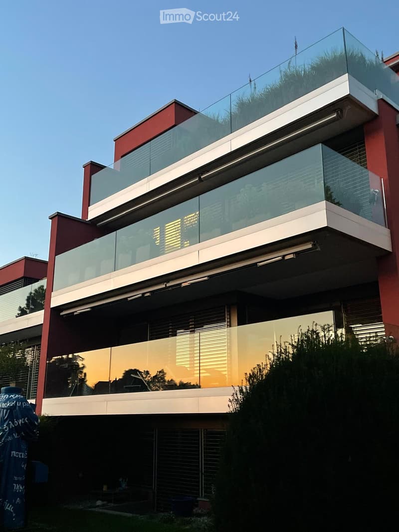 Modern apartment building with glass balconies, red exterior, large windows, visible garden