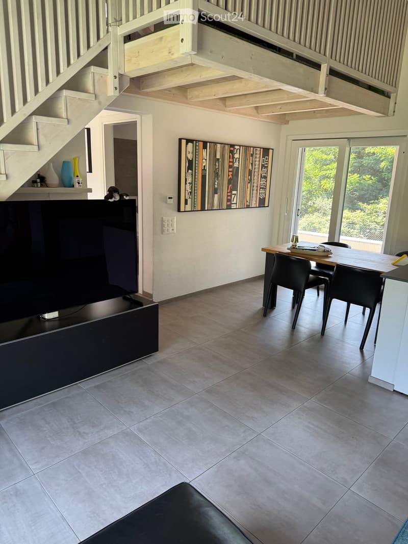 Modern living room with a large TV, sofa, dining table, chairs, and decorative vases. The room features a staircase leading to an upper level and large windows with a view of greenery.
