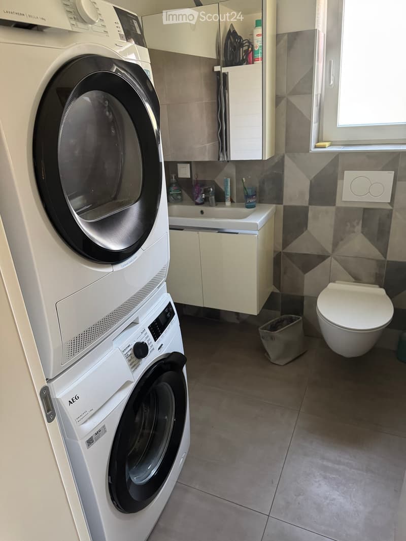 Modern bathroom with a washing machine and dryer stacked, a toilet, a sink with a cabinet, and a small trash bin.