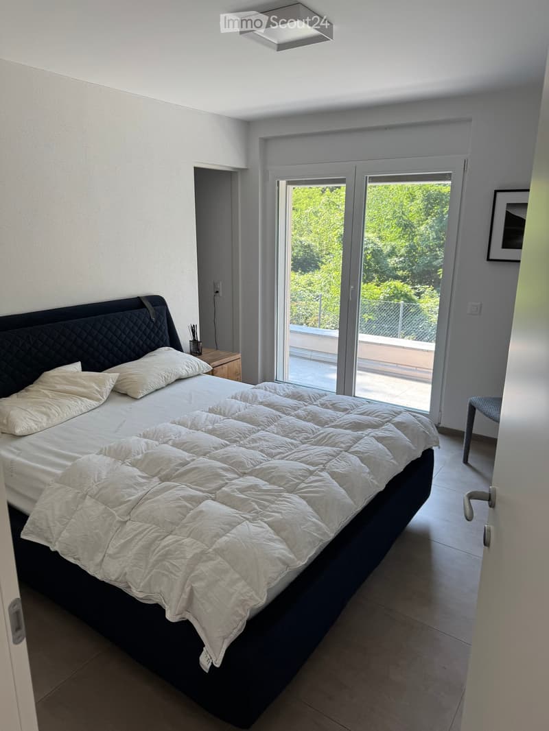 Modern bedroom with a large bed, white bedding, nightstand, and a view through large glass doors to a balcony area.