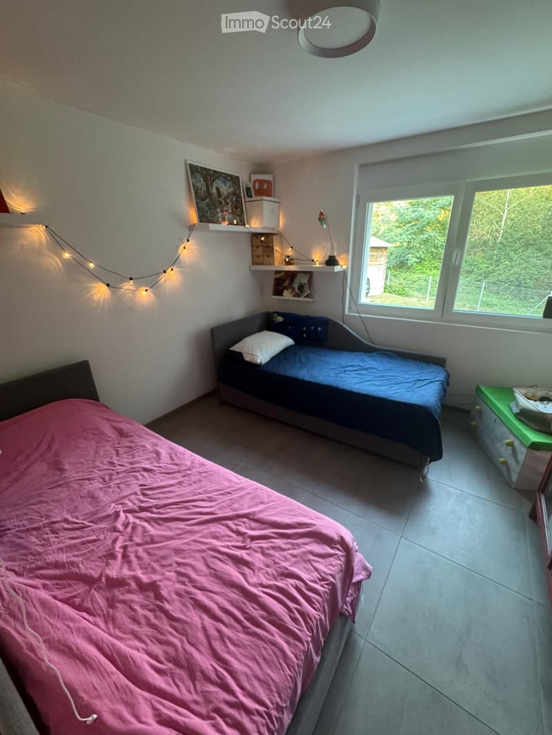 A cozy bedroom with two beds, one with a pink duvet and the other with a blue duvet, a nightstand, shelves with decorative items, and a window with a view of greenery.