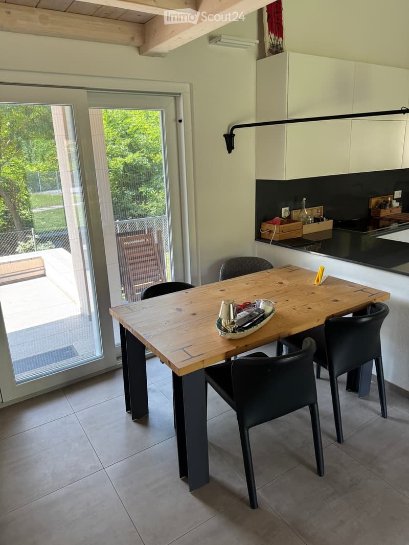Modern kitchen with a wooden dining table, black chairs, and a sleek cabinetry setup. The space features a large window with a view of greenery outside.