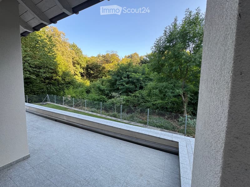 Balcony with tiled floor, metal railing, and a view of a fenced garden area with trees and greenery.