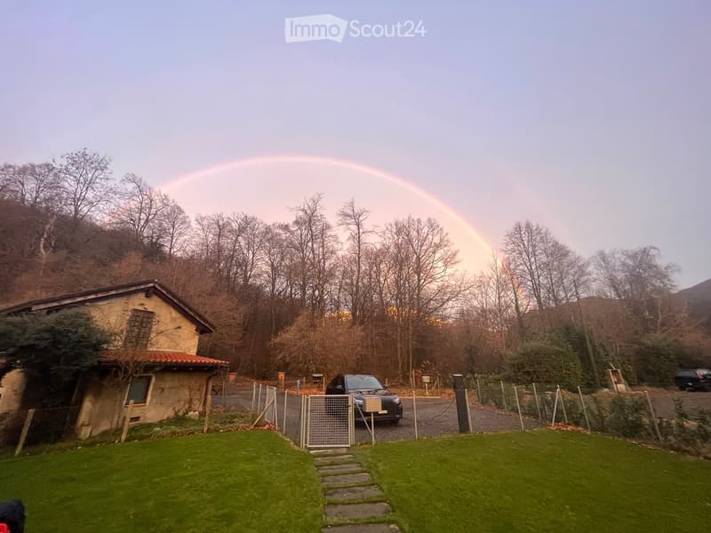 A small, rustic house with a tiled roof and stone walls, surrounded by a grassy lawn and trees, with a car parked nearby and a rainbow visible in the sky.