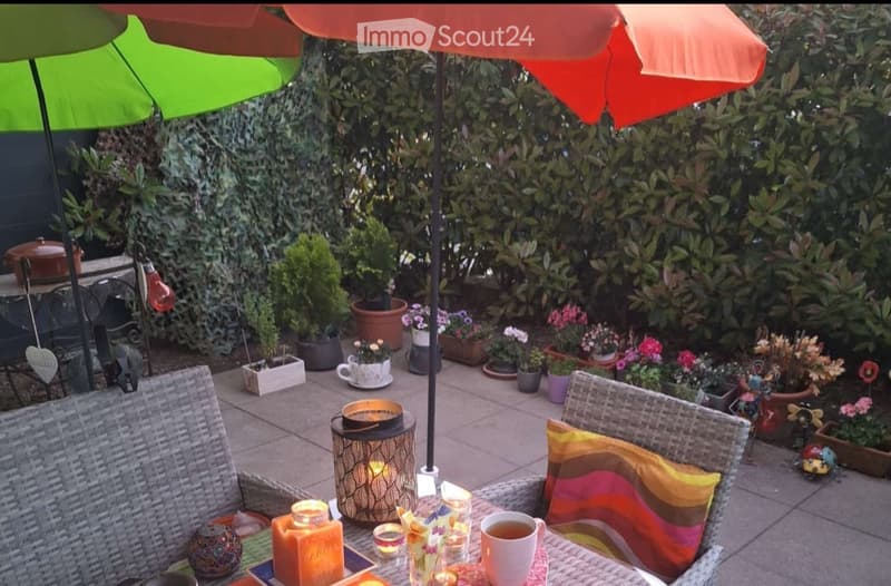A cozy outdoor patio area with wicker chairs, colorful cushions, a table with candles and a teacup, surrounded by potted plants and flowers, with colorful umbrellas providing shade.