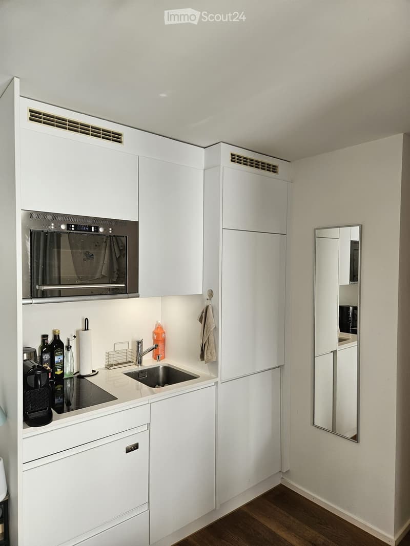 Modern kitchen with white cabinets, stainless steel appliances, and a small sink area. Includes a coffee maker, bottles, and a paper towel holder.