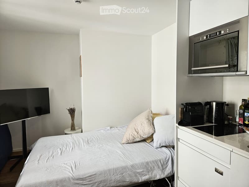 A compact studio apartment with a bed, television, small table with a vase, and a kitchenette area with an oven, coffee maker, and kettle.
